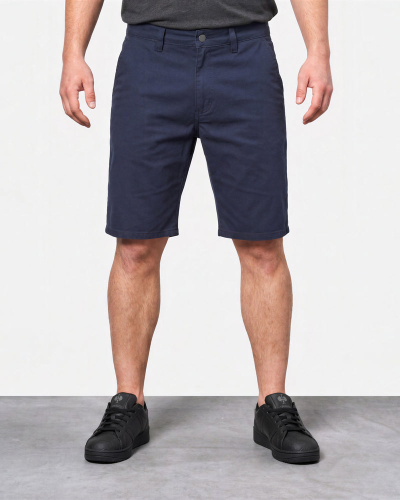 A person wearing navy blue knee-length shorts, a dark gray t-shirt, and black sneakers, standing against a white wall and gray floor.
