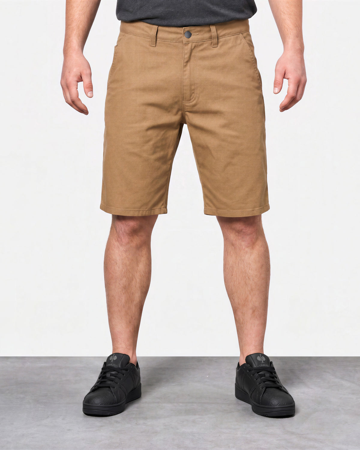 A person wearing tan chino-style shorts, shown from the waist down, standing against a light gray studio background with a gray floor. The shorts are the product being featured, paired with a dark gray shirt and black sneakers.