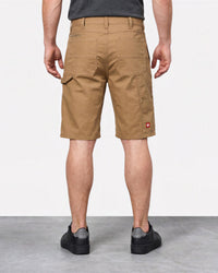 A man viewed from the back wearing tan cargo shorts, a dark gray t-shirt, and black slip-on shoes, standing against a white studio wall.
