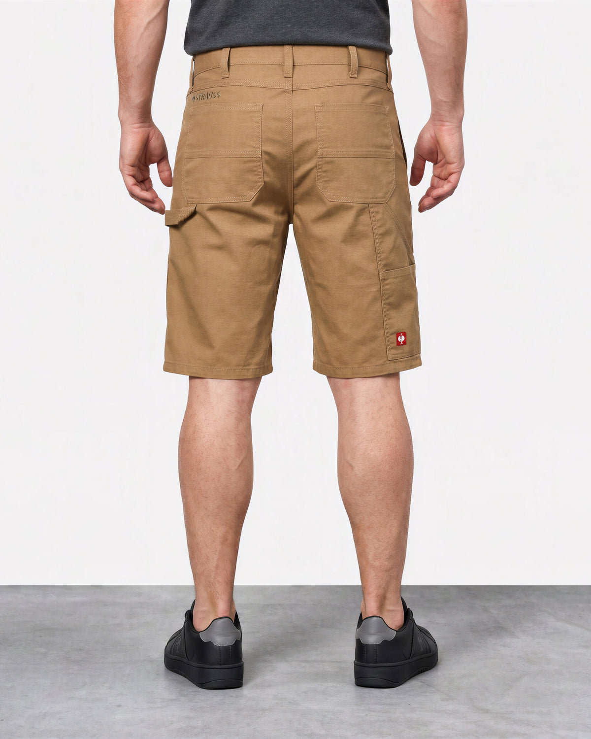 A man viewed from the back wearing tan cargo shorts, a dark gray t-shirt, and black slip-on shoes, standing against a white studio wall.