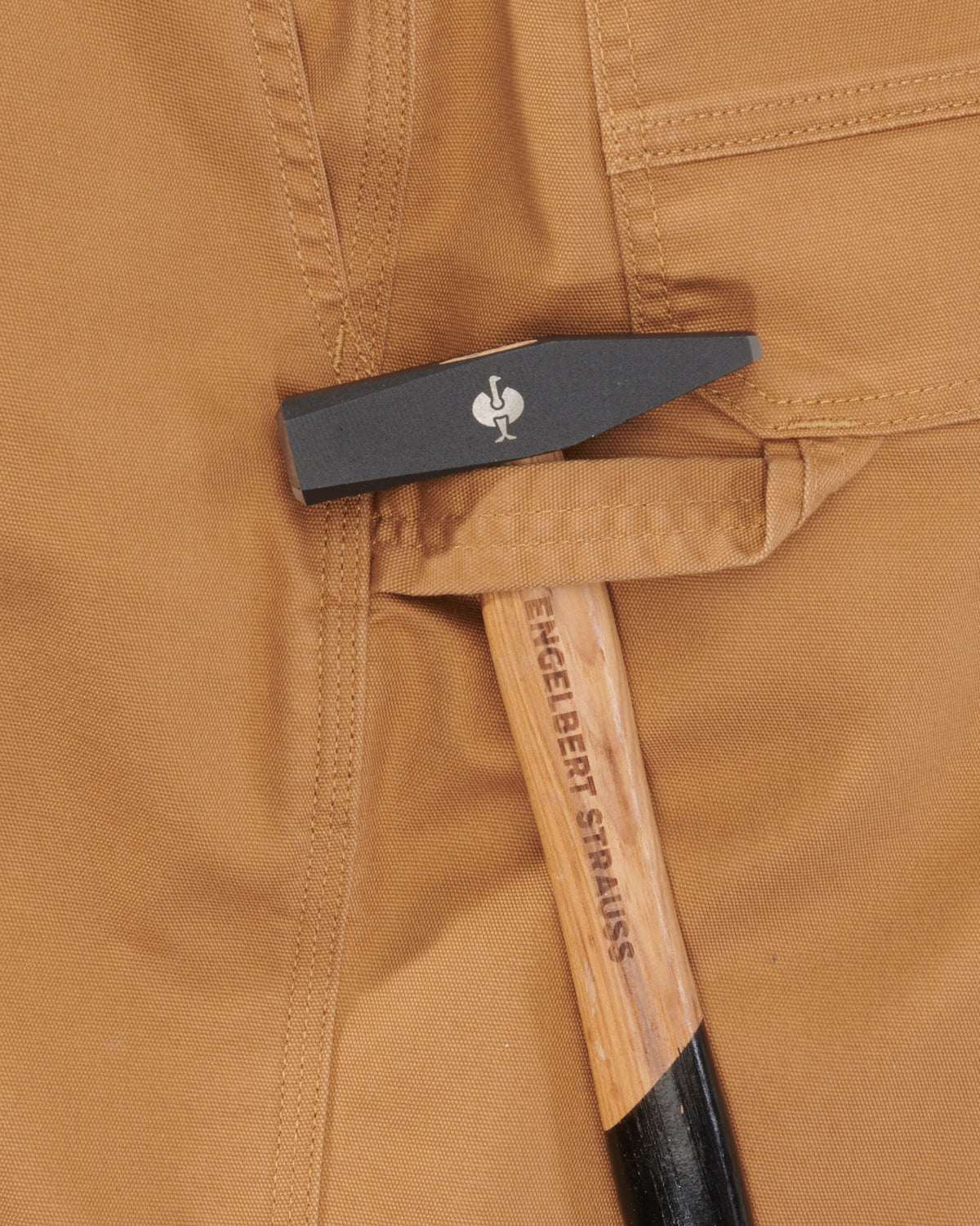 Close-up of a tan workwear jacket with a black and silver pocket knife tucked into the fabric. The knife has a white rooster logo, and the handle shows the brand name 'ENGELBERT STRAUSS'.