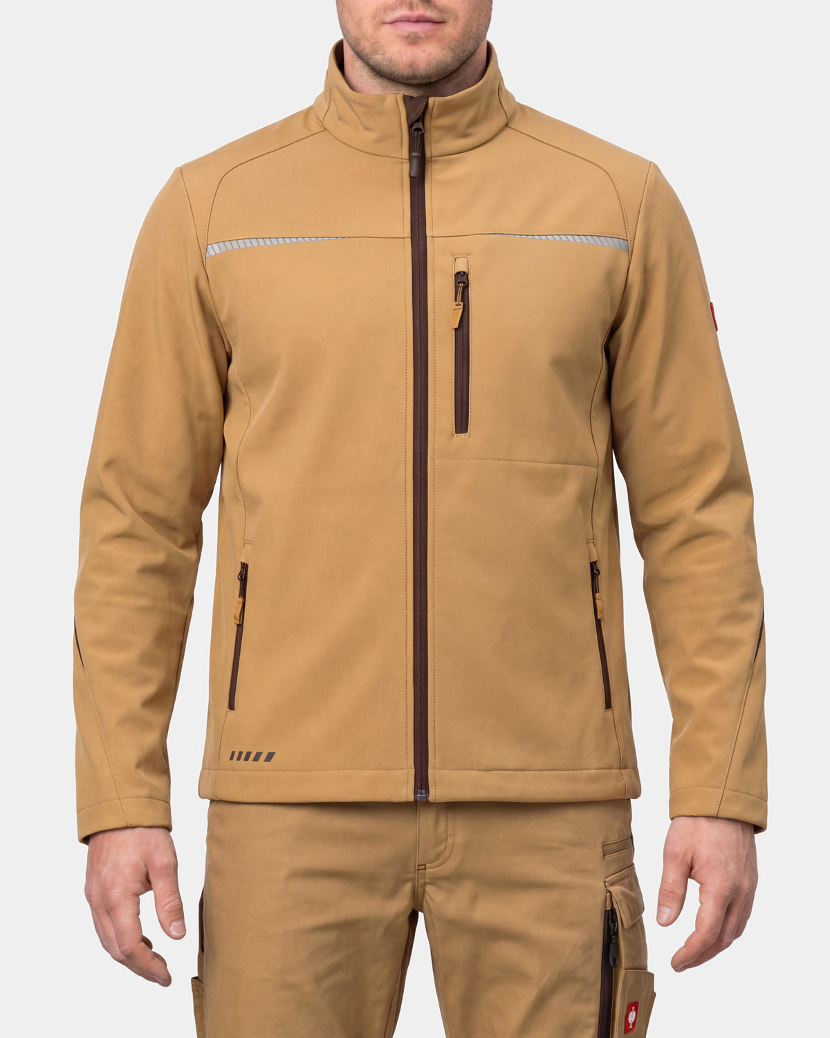 A man wearing a tan softshell jacket with a high collar, front zipper, and zippered pockets, paired with matching tan pants, against a light gray background.