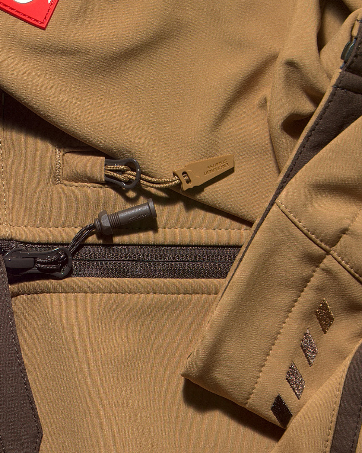 Close-up of a tan technical jacket showing a zippered pocket with a tan zipper pull, dark zipper track, and a red square logo patch on the upper left.