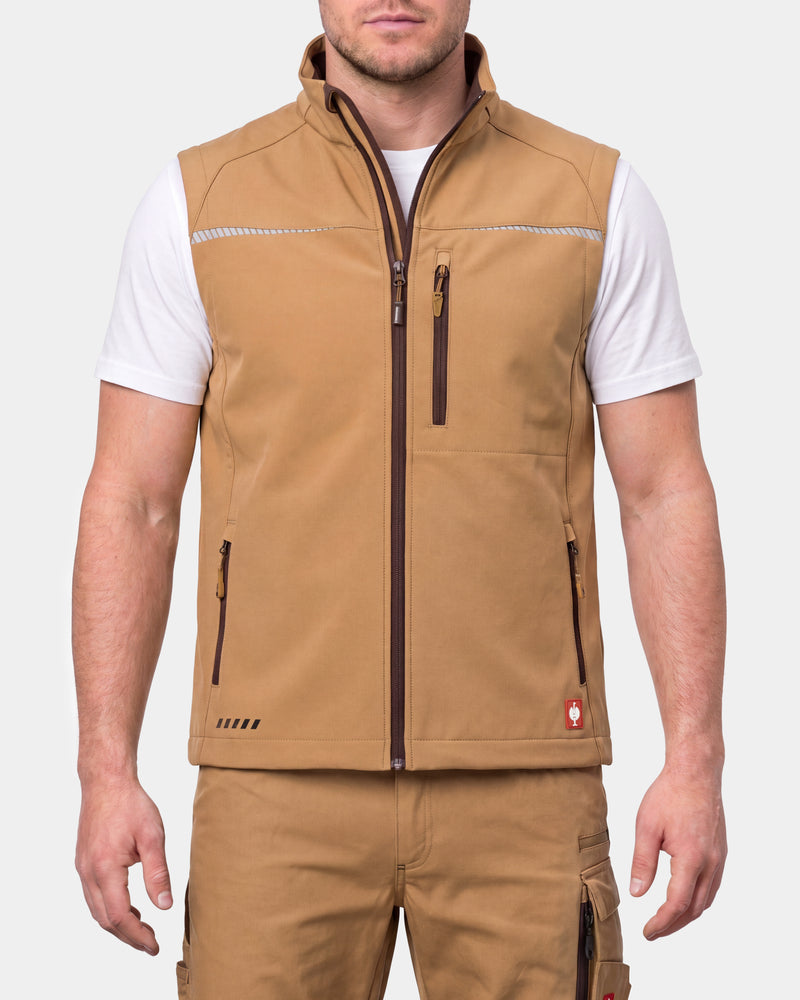 A man wearing a tan zip-up vest over a white t-shirt and matching tan cargo pants, shown from the neck down against a light gray studio background.