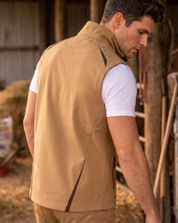 A man wearing a tan insulated vest over a white t-shirt, standing in a rustic barn setting.