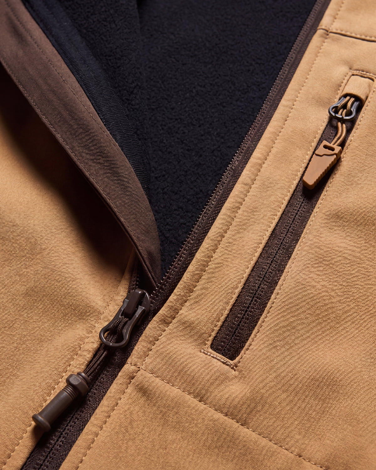 Close-up of a tan fleece-lined zippered jacket showing the tan exterior fabric, dark brown zipper trim, and a black fleece interior.