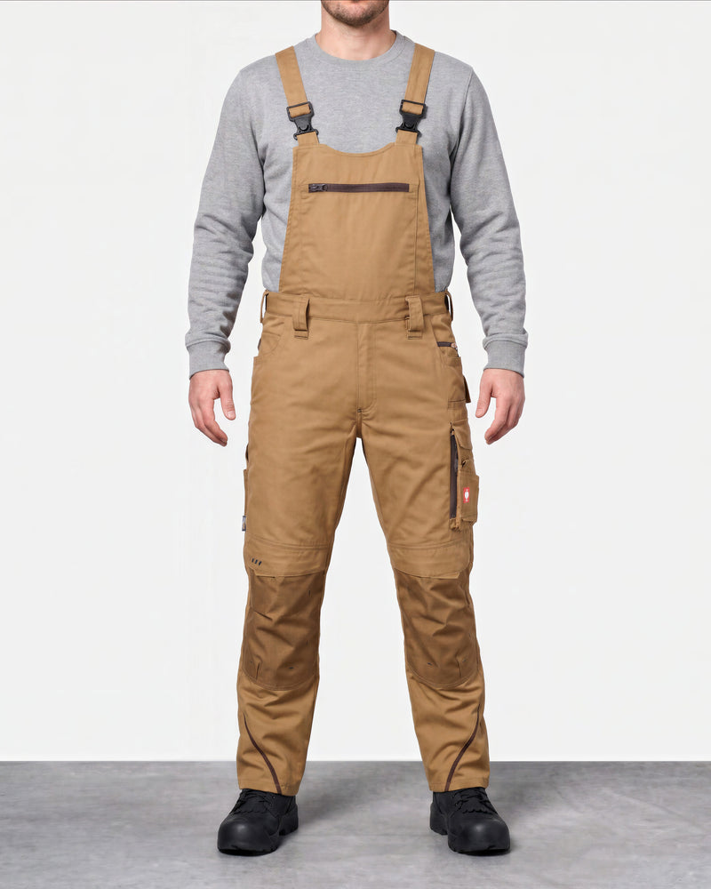 A man wearing tan work bib overalls over a gray long-sleeve shirt, standing against a light gray studio background.