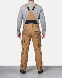 A man viewed from the back wearing tan cargo-style bib overalls with black shoulder straps and a black waist band, layered over a heather gray long-sleeve shirt. The overalls have black piping details along the legs.