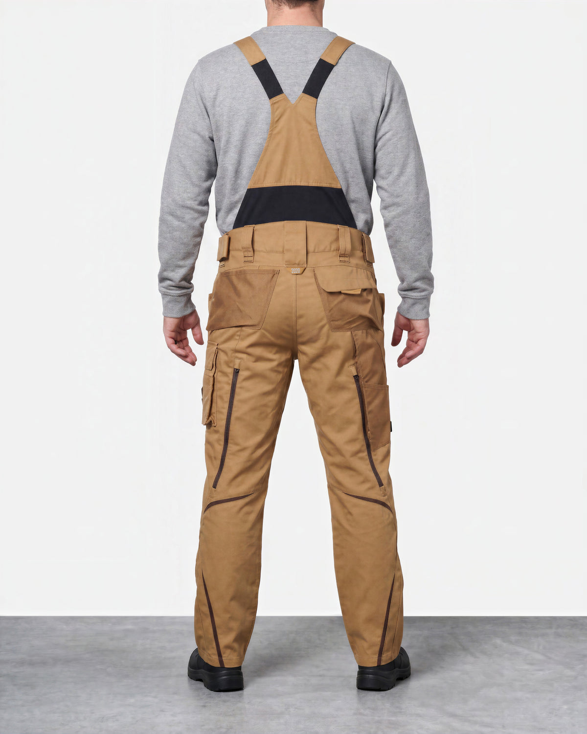 A man viewed from the back wearing tan cargo-style bib overalls with black shoulder straps and a black waist band, layered over a heather gray long-sleeve shirt. The overalls have black piping details along the legs.