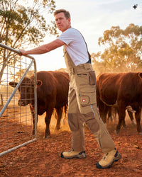 A man wearing tan agricultural overalls, a white t-shirt, and work boots stands in a dusty farmyard at sunset, leaning on a metal gate with cows in the background.