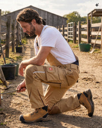 A man kneels in a dirt farmyard wearing tan work overalls over a white t-shirt, with brown work boots and a small red logo patch on the thigh pocket.