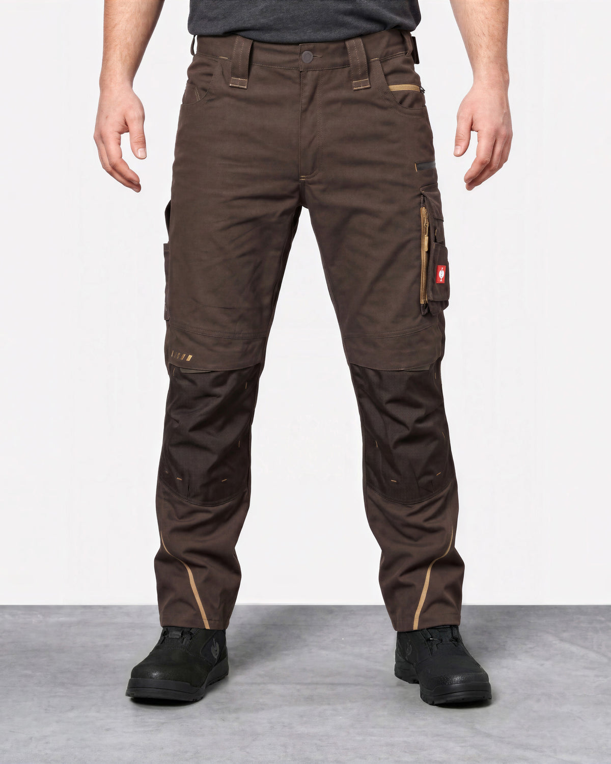 A person wearing dark brown work pants with reinforced knee panels and black work boots, standing against a light gray studio background.