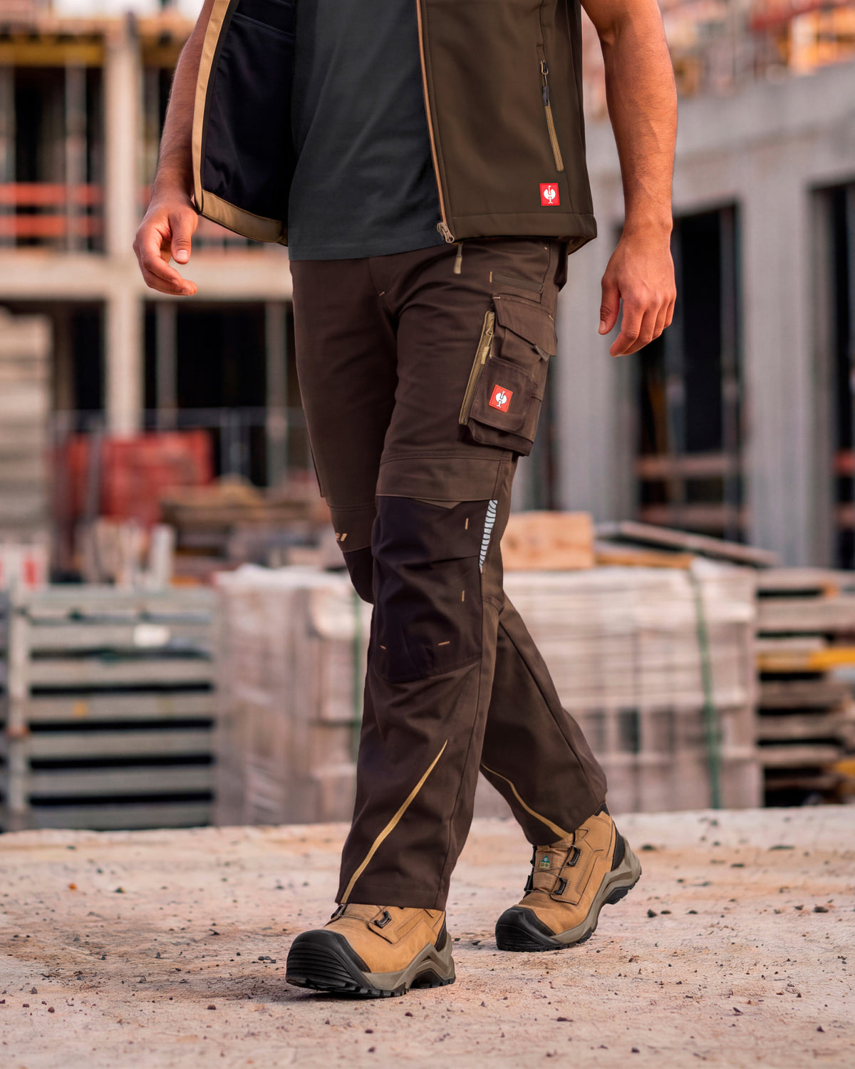 A man wearing dark brown and black technical work pants with utility pockets, a dark vest, and work boots, walking on a concrete surface at a construction site.