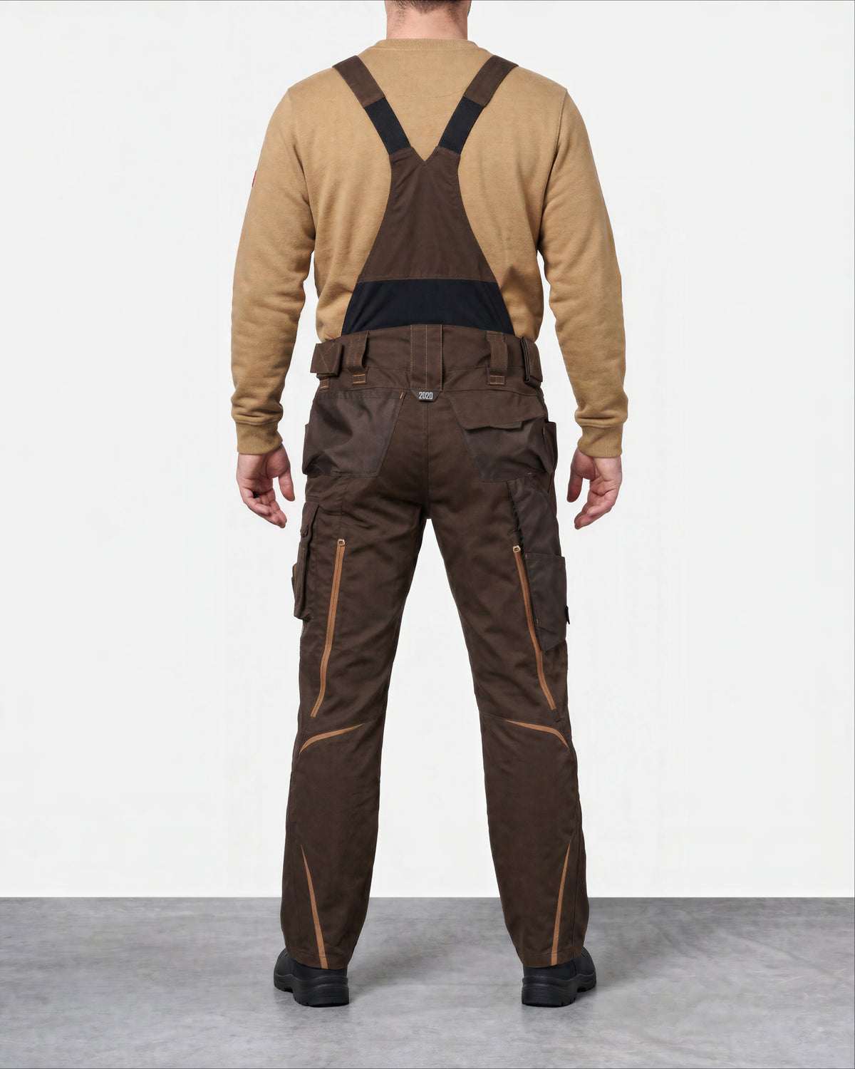 A man viewed from the back wearing dark brown work bib overalls with black shoulder straps and a tan long-sleeve shirt, standing against a light gray studio background.