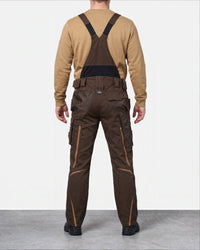 A man viewed from the back wearing dark brown work bib overalls with black shoulder straps and a tan long-sleeve shirt, standing against a light gray studio background.