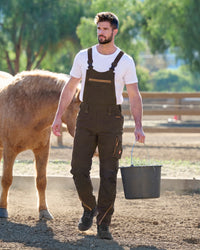A man wearing dark brown work overalls over a white t-shirt walks in a dusty paddock while holding a bucket of grass. A light brown horse is visible on the left, with trees and a fence in the background.