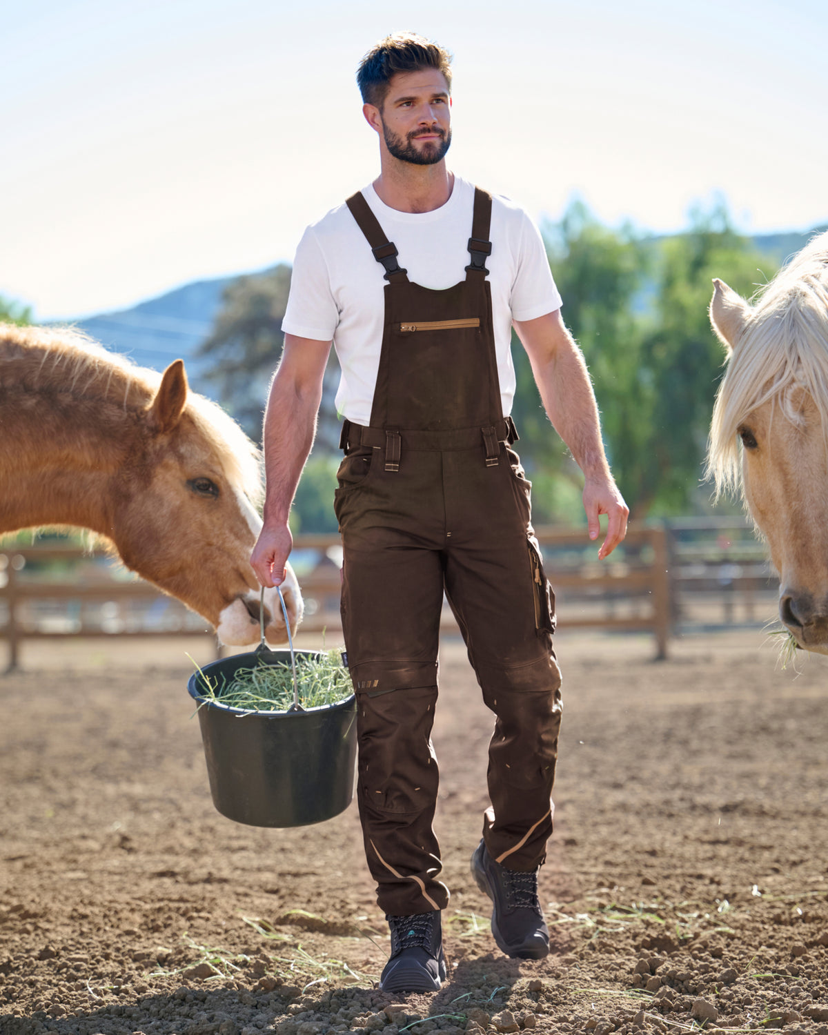 A man wearing dark brown overalls over a white t-shirt walks in a horse paddock while holding a bucket of hay. Two horses are visible on either side of him.