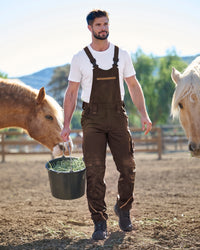 A man wearing dark brown overalls over a white t-shirt walks in a horse paddock while holding a bucket of hay. Two horses are visible on either side of him.