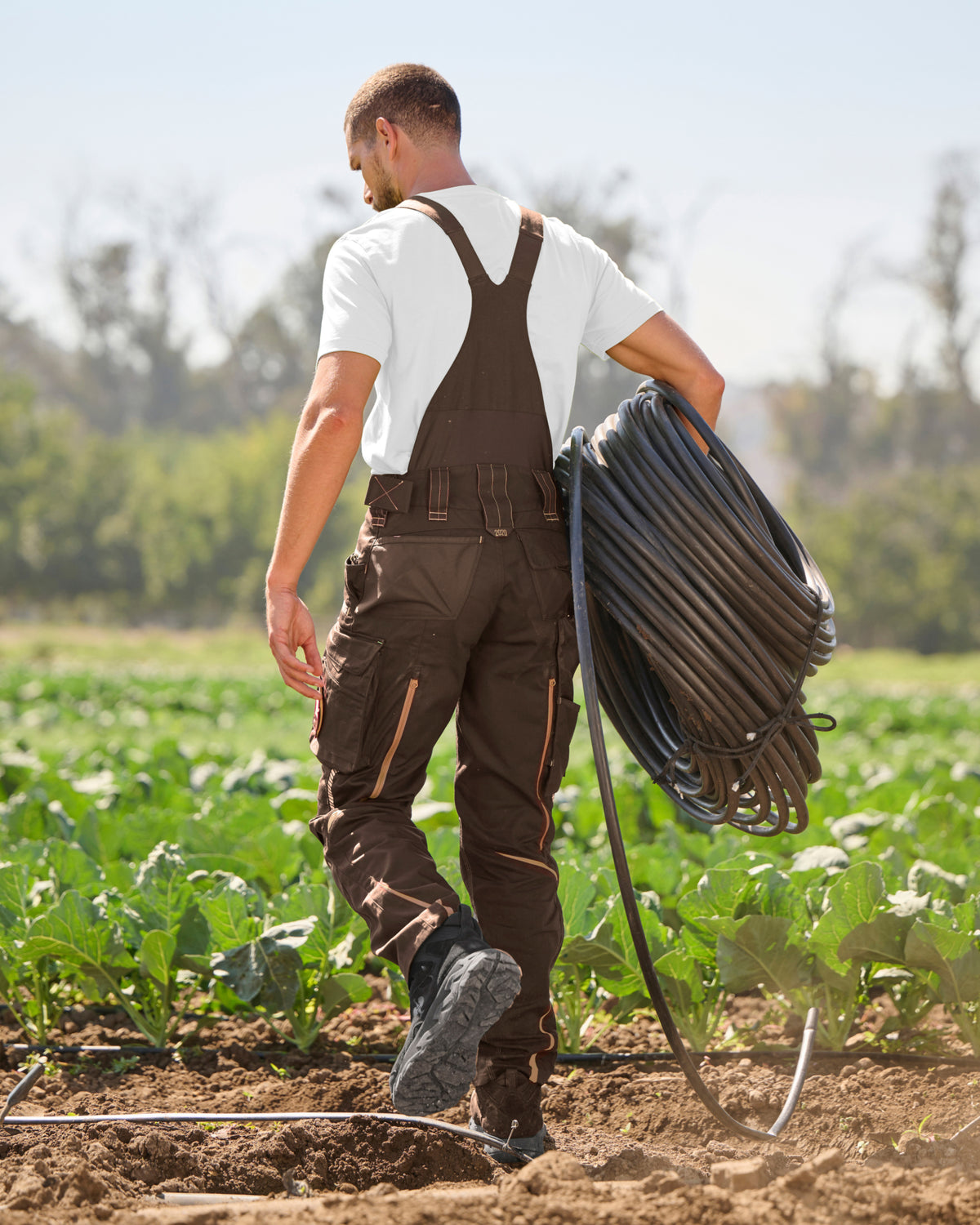 A man in brown work overalls and a white t-shirt walks through a farm field while carrying a large coil of black irrigation tubing over his shoulder.