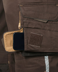Close-up of a brown tactical utility pocket on a bib, showing a beige interior slip pocket with a black smartphone inserted and a brown hook-and-loop patch on the front.