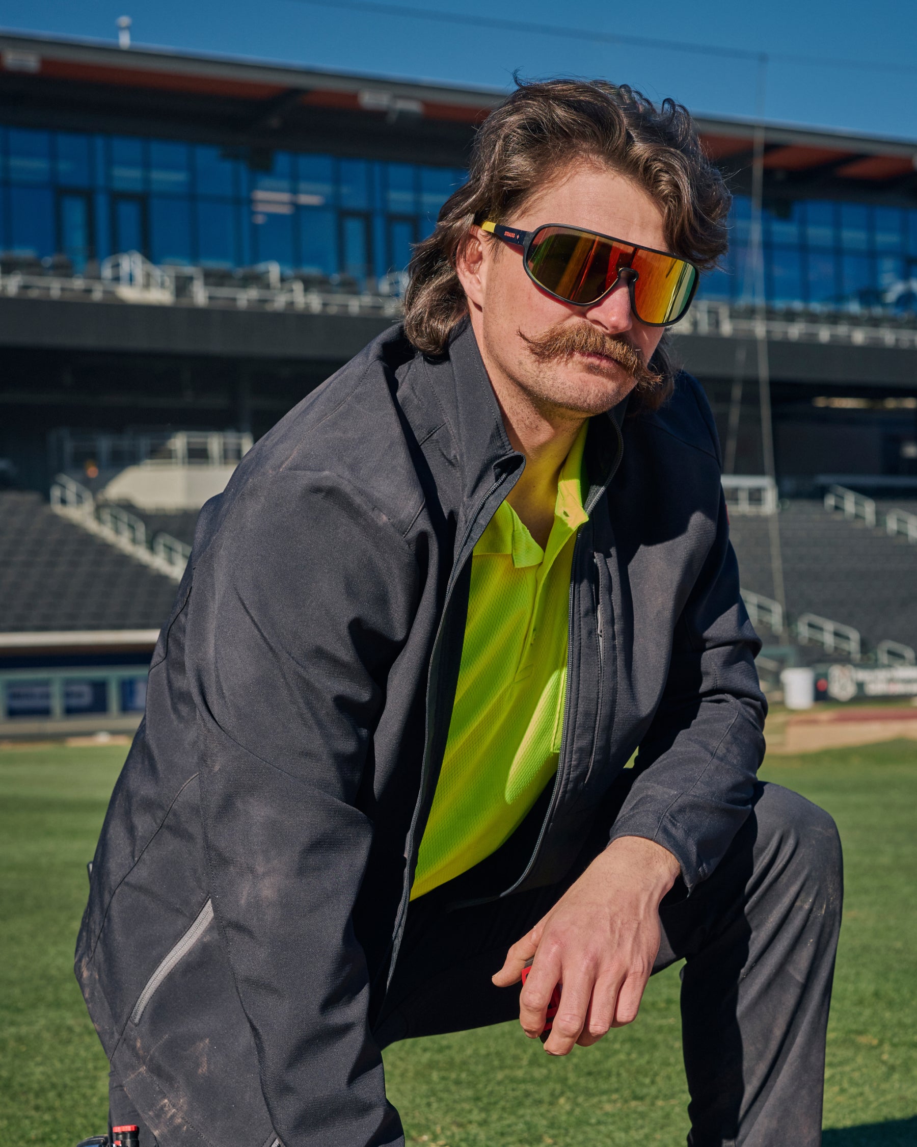 A person wears black and high-vis yellow STRAUSS e.s.ambition race sunglasses with a utility jacket in an outdoor work setting.