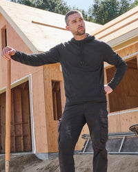 A man wearing a black long-sleeve hoodie and black cargo pants stands in front of a wooden house under construction, holding a shovel.