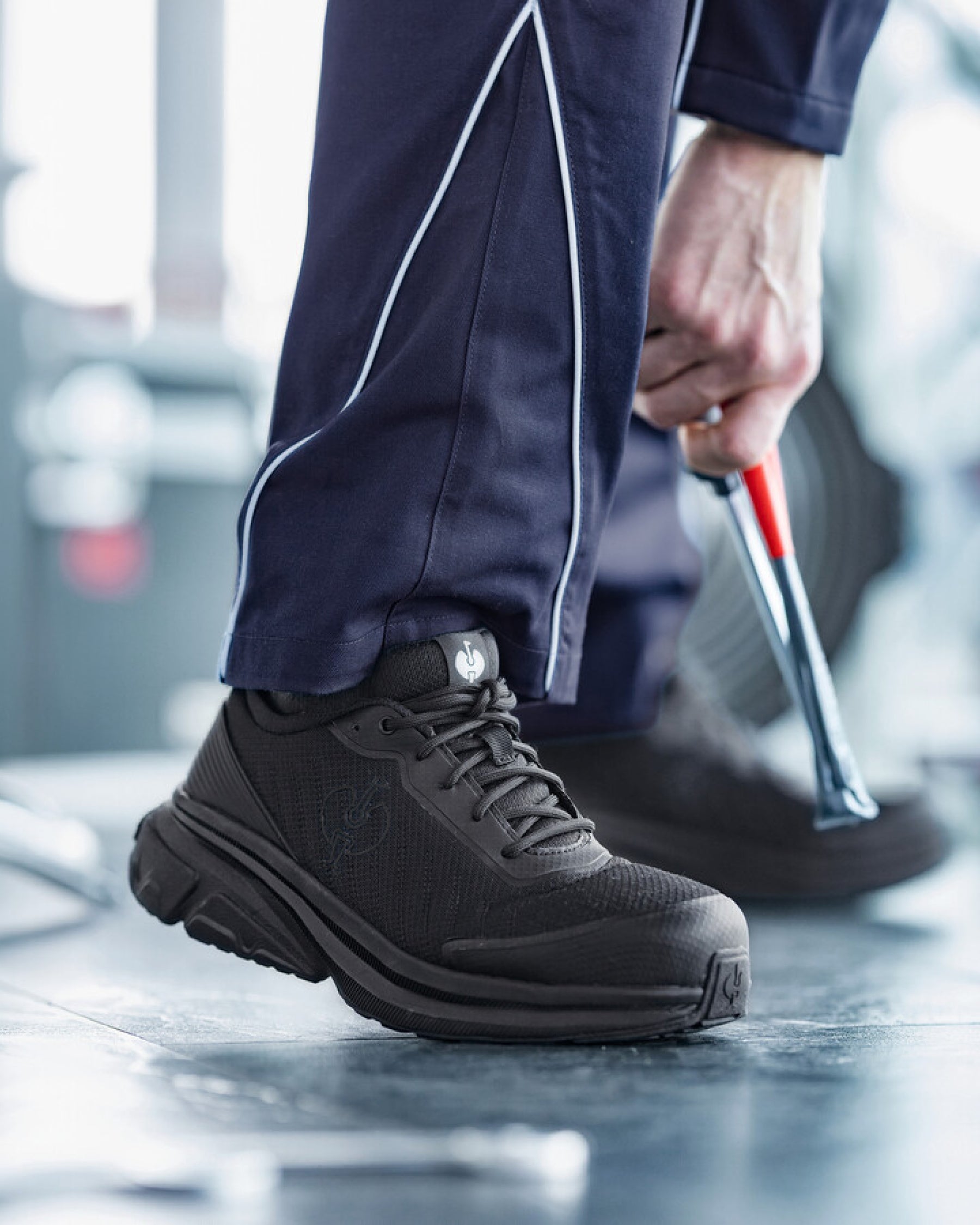 A close-up of a person wearing dark gray and black athletic shoes with navy blue pants featuring white piping, standing on a gym floor.