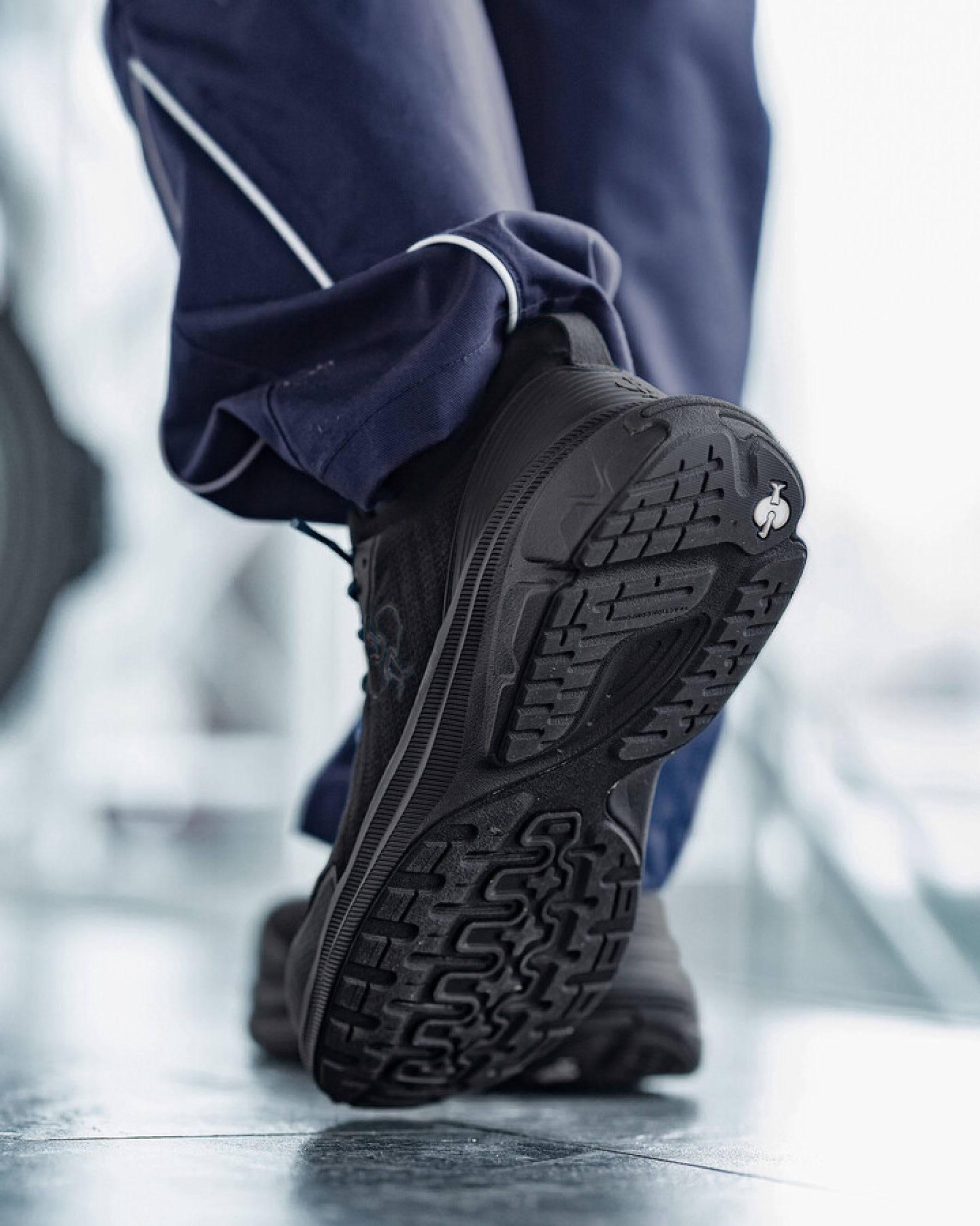 A close-up shot of a person's foot wearing a black athletic sneaker, showing the textured black outsole, with navy blue pants featuring white piping visible in the background.