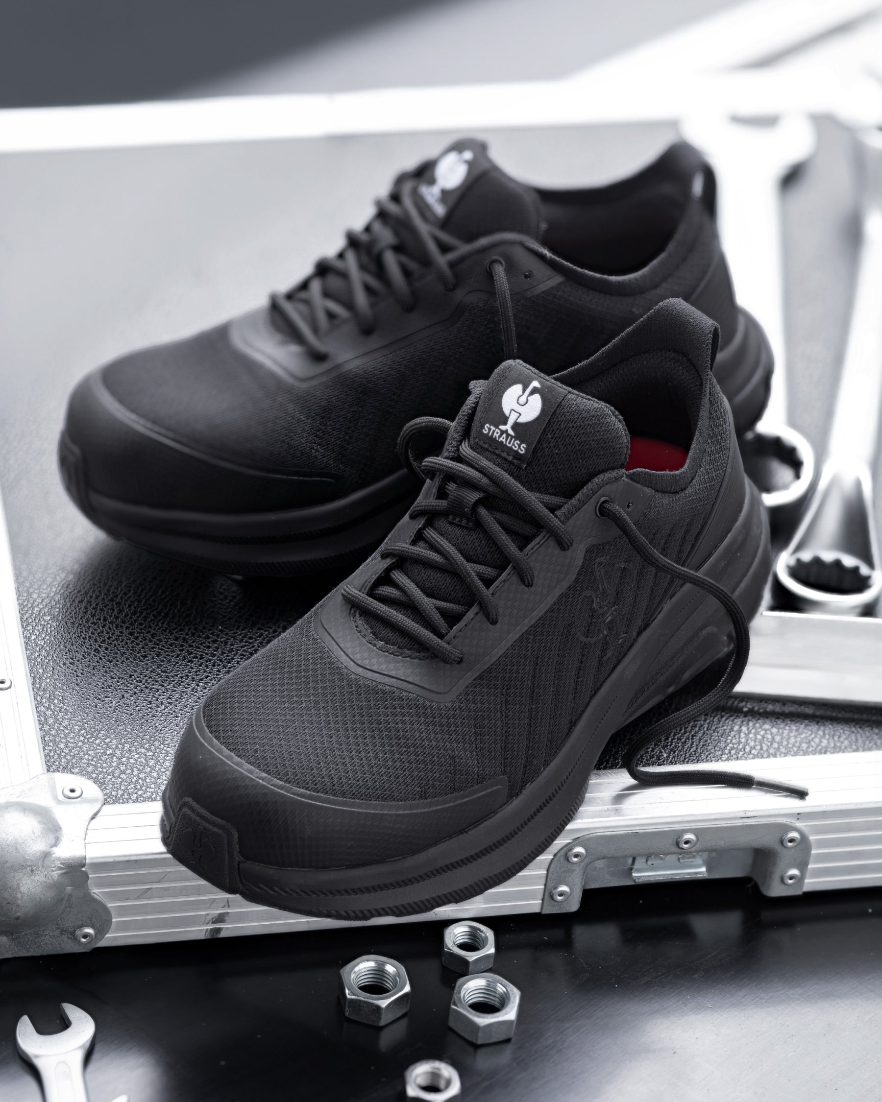 A pair of black work sneakers resting on the corner of a silver metal equipment case, with wrenches and nuts nearby on a dark surface.