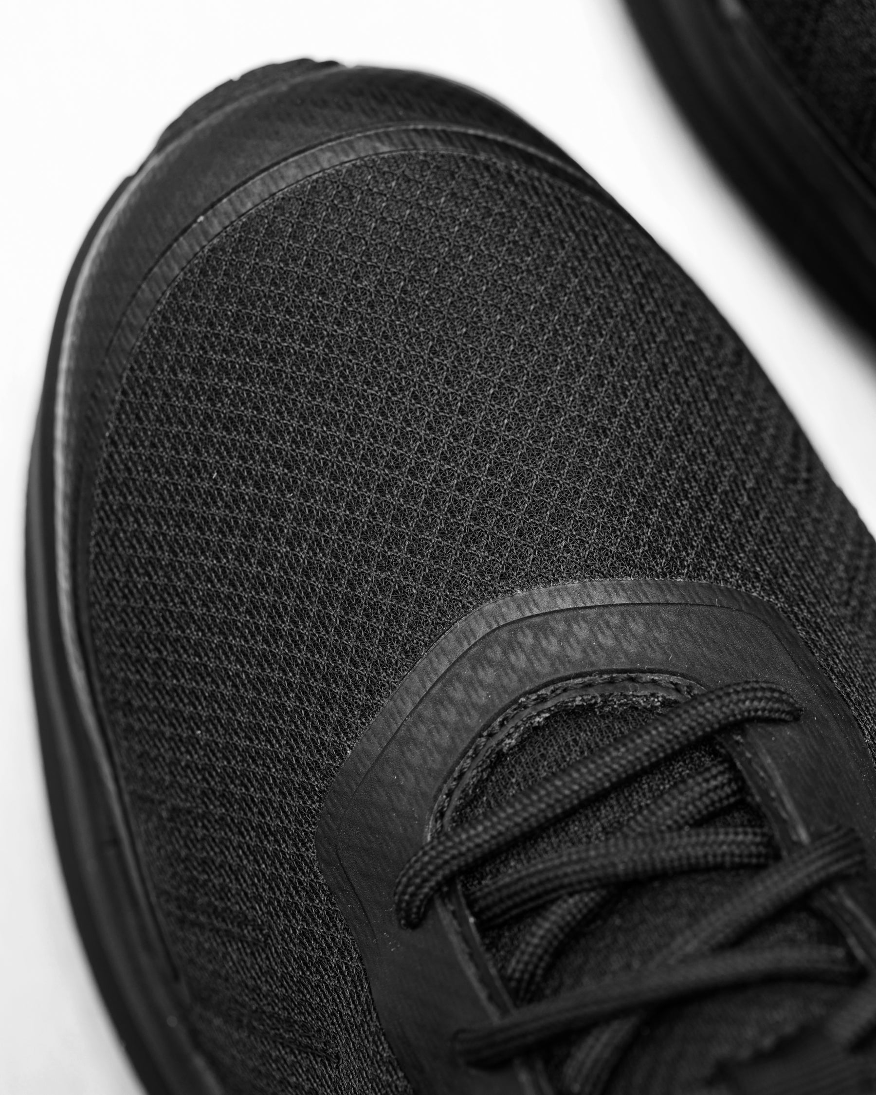 Close-up of a black athletic shoe showing the textured mesh upper, laces, and a portion of the sole on a white background.
