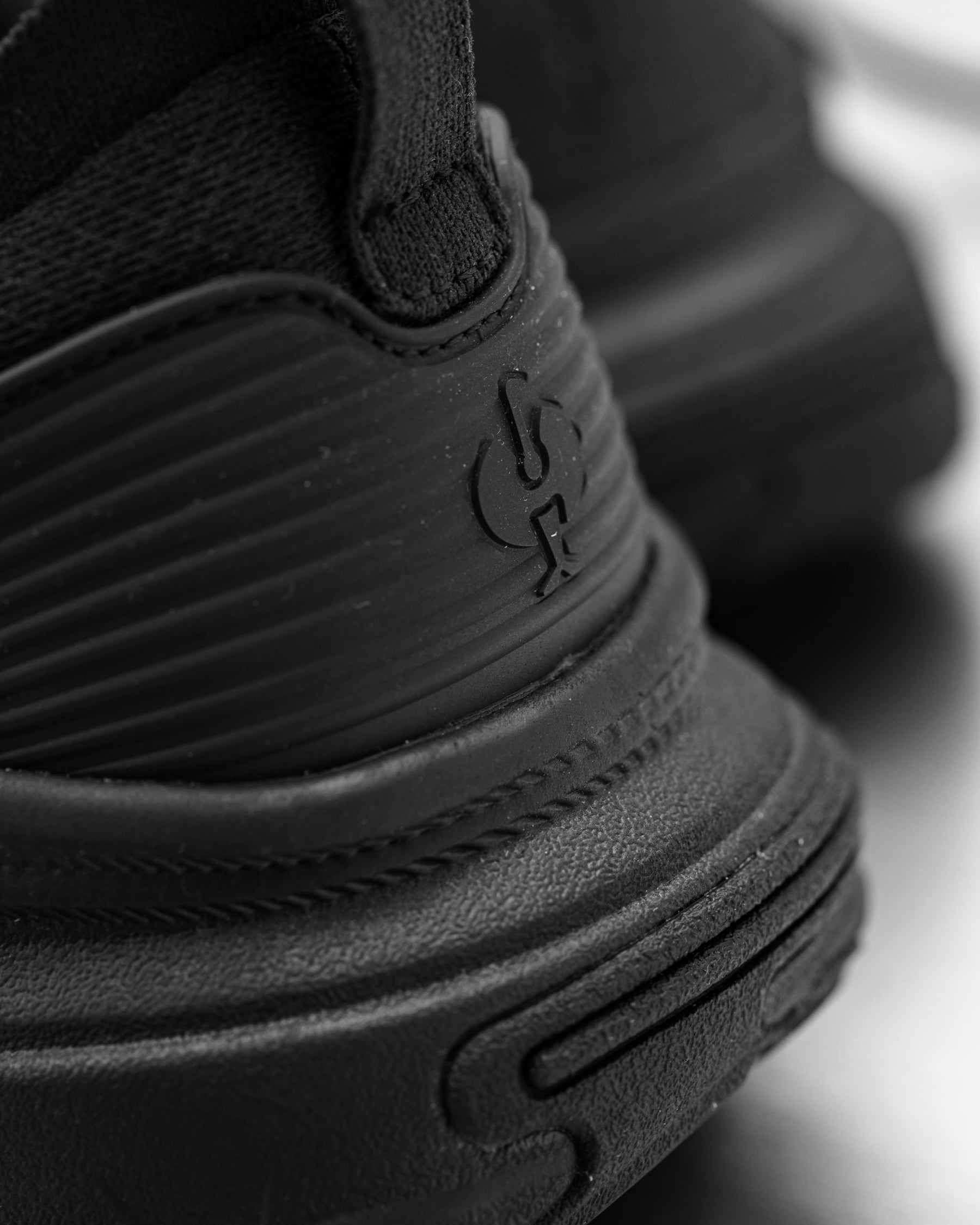 Close-up of a black sneaker showing the textured rubber sole, stitched side panel, and embossed logo.