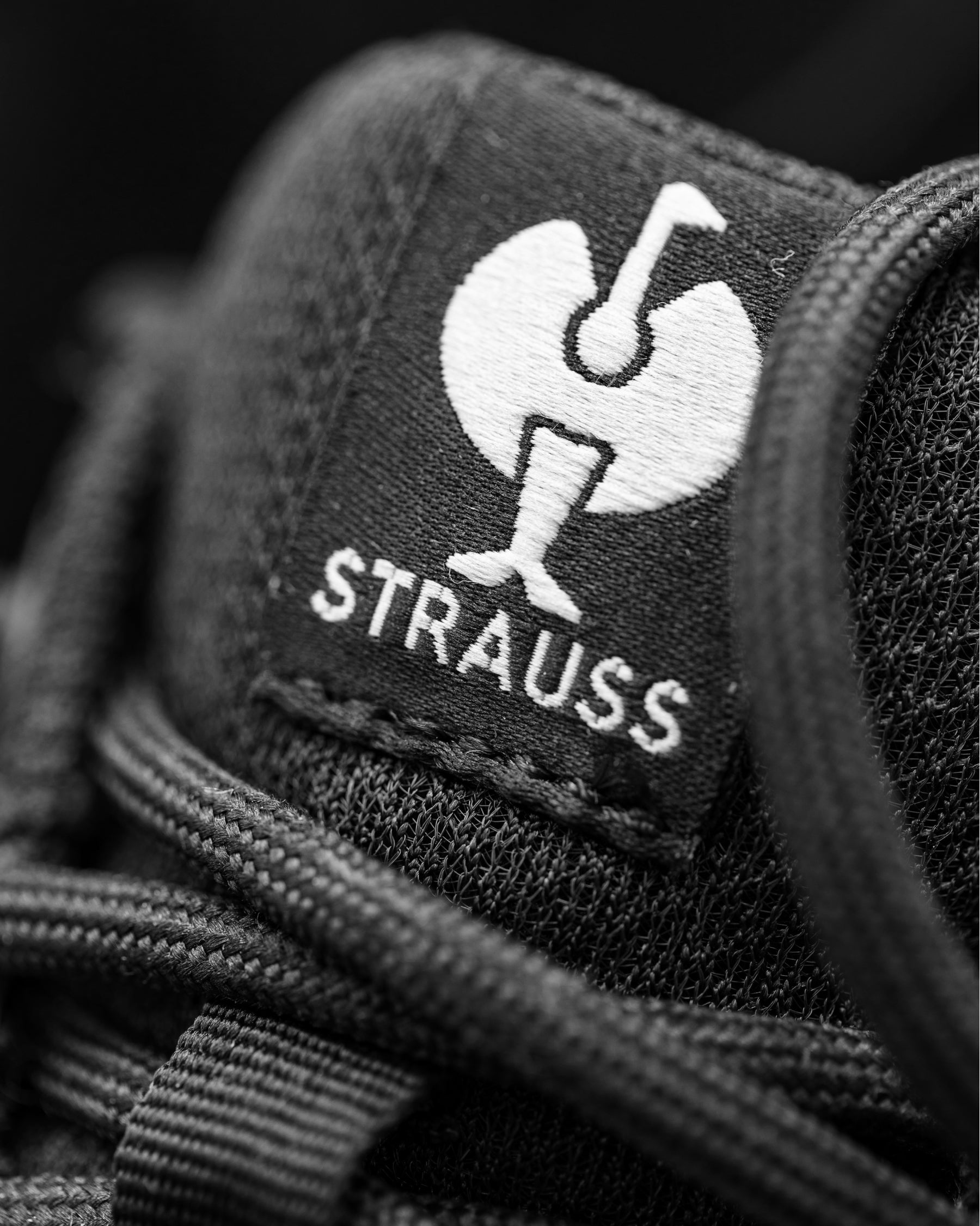 Close-up of a black shoe tongue patch with a white embroidered Strauss logo and the word 'Strauss' in white text.