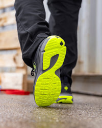 A close-up of a person's leg walking, showing the outsole of a black and neon yellow-green running shoe. The shoe has a black upper with a bright neon yellow-green treaded sole and matching neon yellow-green accents.