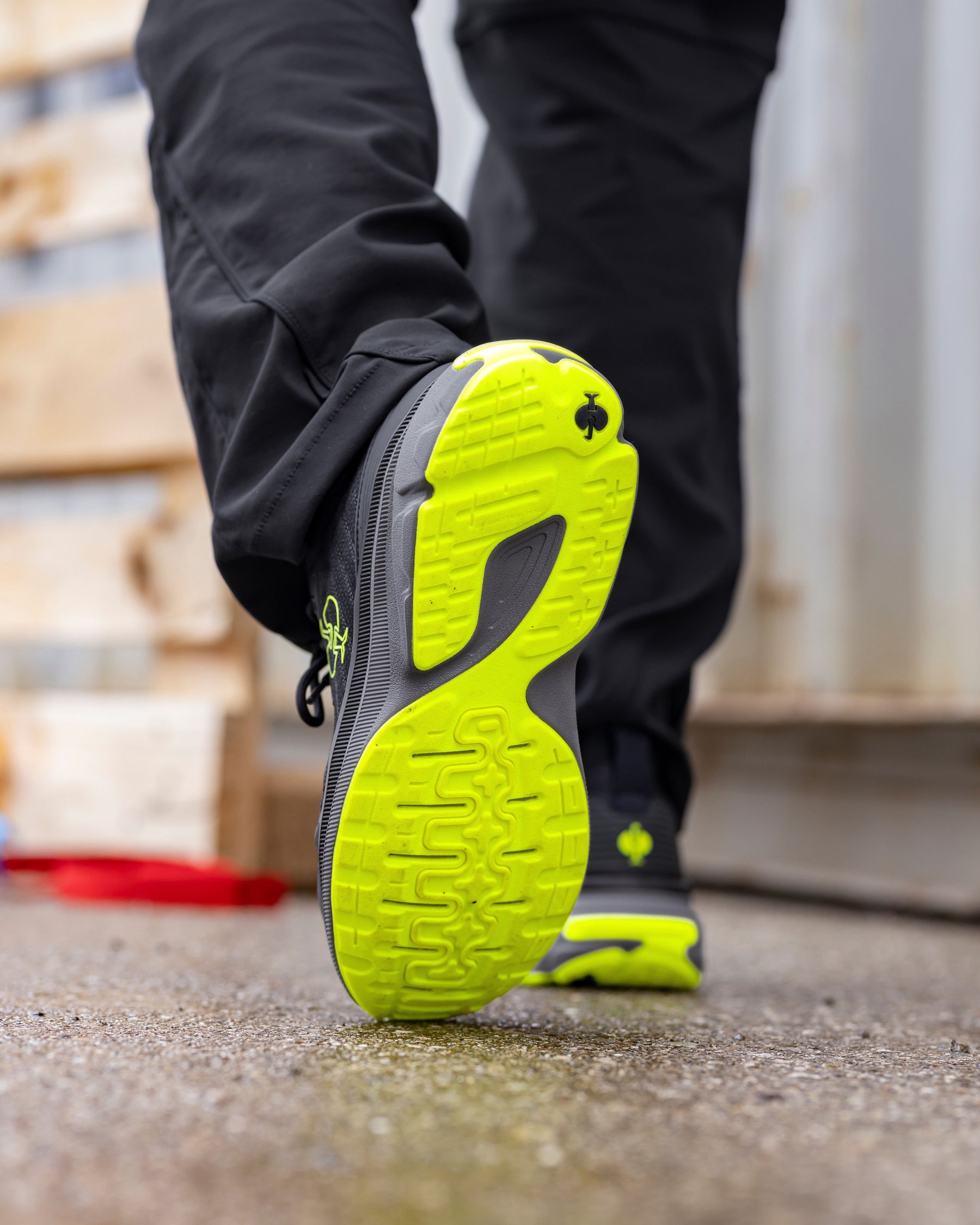 A close-up of a person's leg walking, showing the outsole of a black and neon yellow-green running shoe. The shoe has a black upper with a bright neon yellow-green treaded sole and matching neon yellow-green accents.