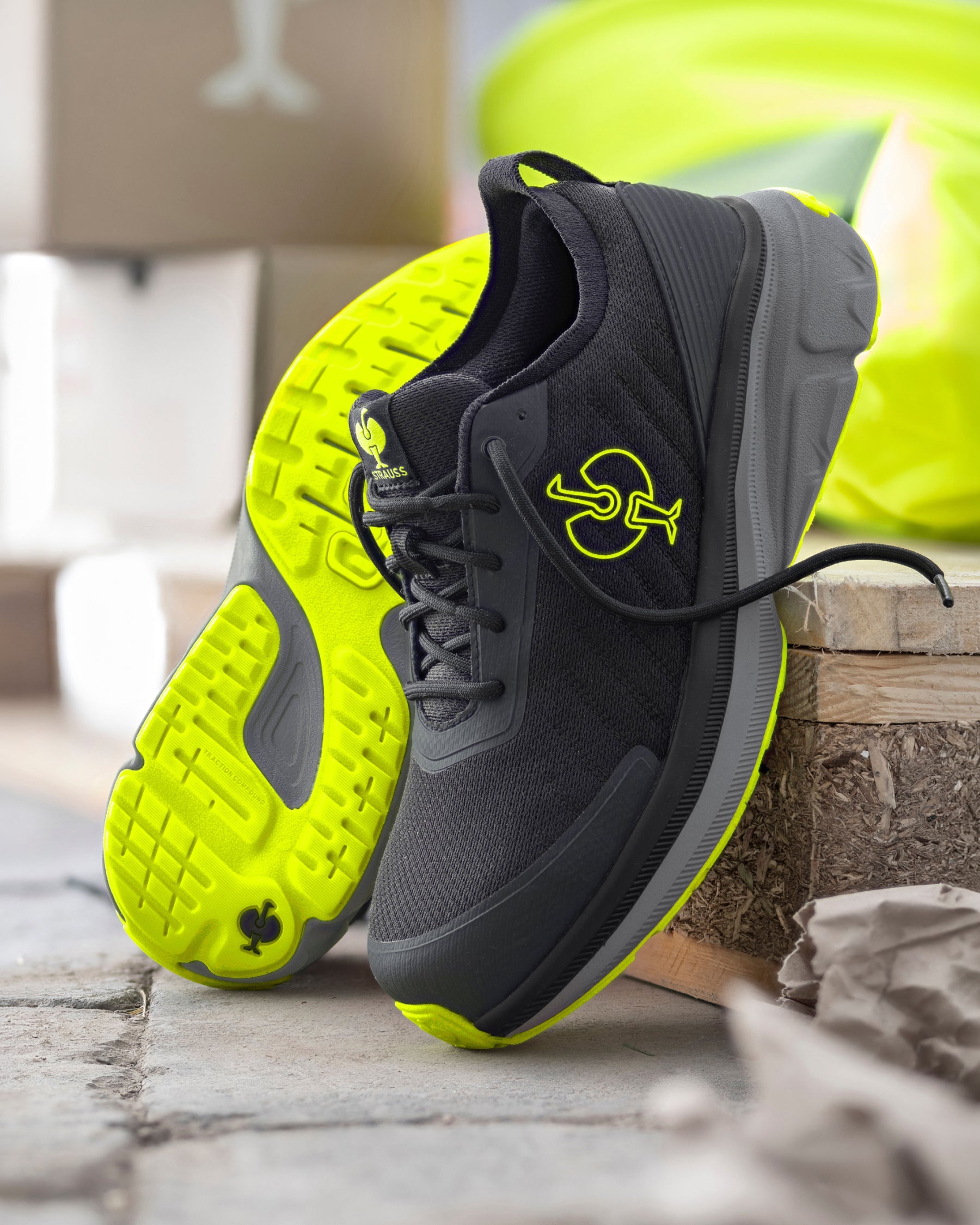 A pair of athletic sneakers in black and neon yellow-green, with one shoe standing upright and the other lying on its side to show the sole. The shoes are styled on a rustic wooden block over a stone surface.