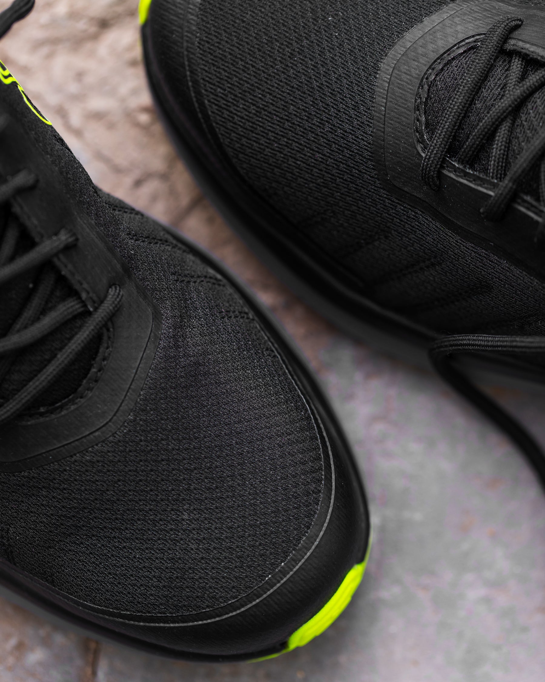 Close-up of a black athletic shoe showing the textured mesh upper, black laces, and a bright neon yellow-green accent on the toe bumper, resting on a gray stone surface.