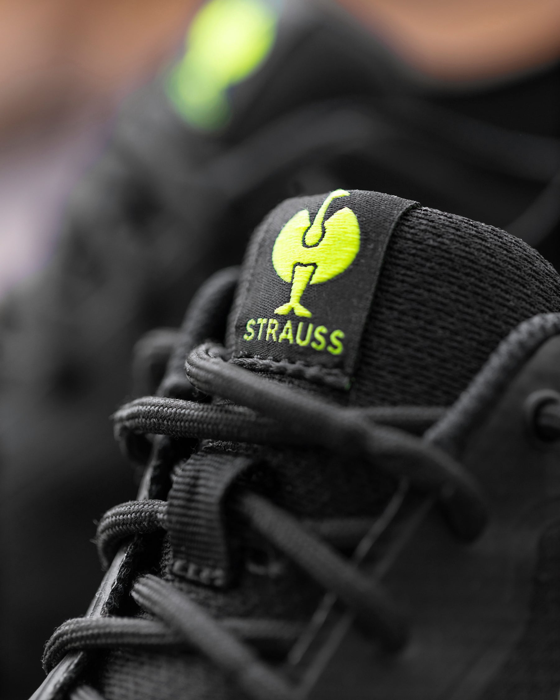 Close-up of a black sneaker with a black tongue label featuring a bright neon yellow-green Strauss logo and the word 'STRAUSS' in matching neon yellow-green.