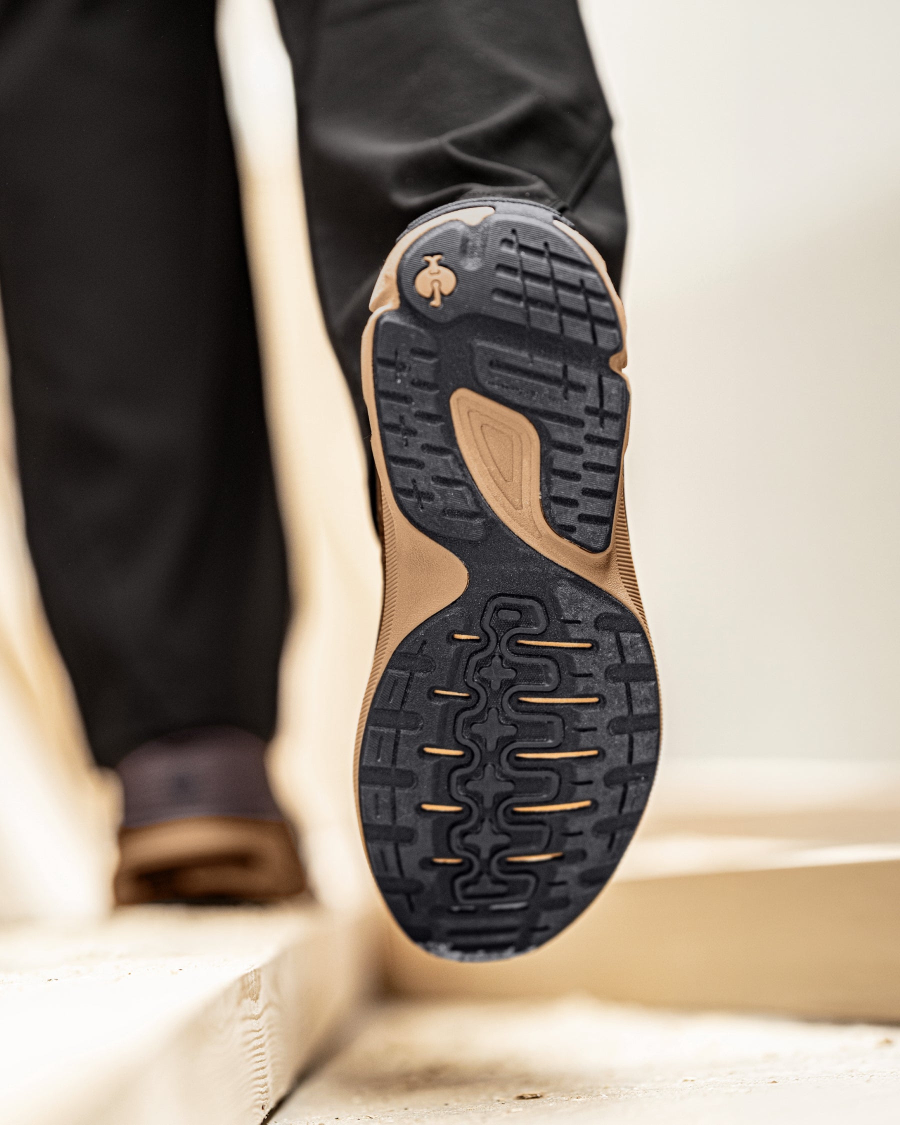 Close-up of the outsole of a hiking shoe being worn, showing a black treaded rubber bottom with tan accents and a small tree logo, with a person's legs in dark pants walking on stone steps.