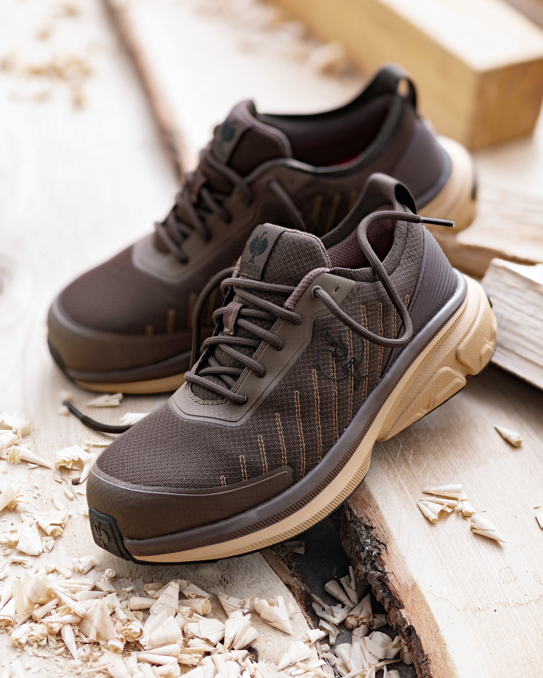 A pair of dark brown athletic sneakers with beige midsoles, styled on a rustic wooden surface with wood shavings.