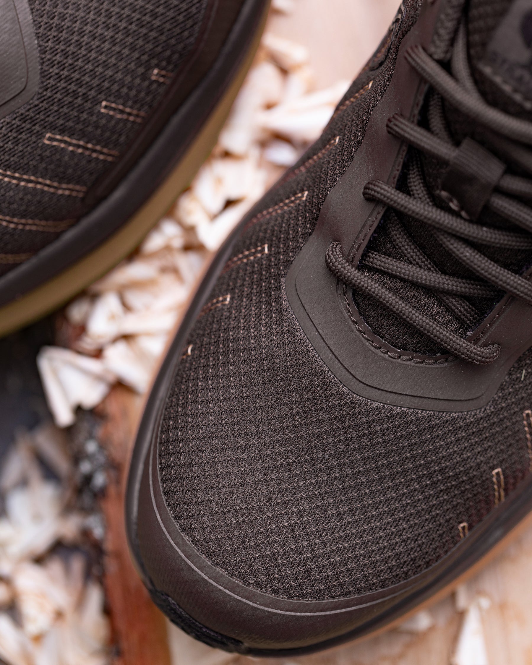 Close-up of a dark brown mesh athletic shoe with matching laces, shown against a background of wood shavings.