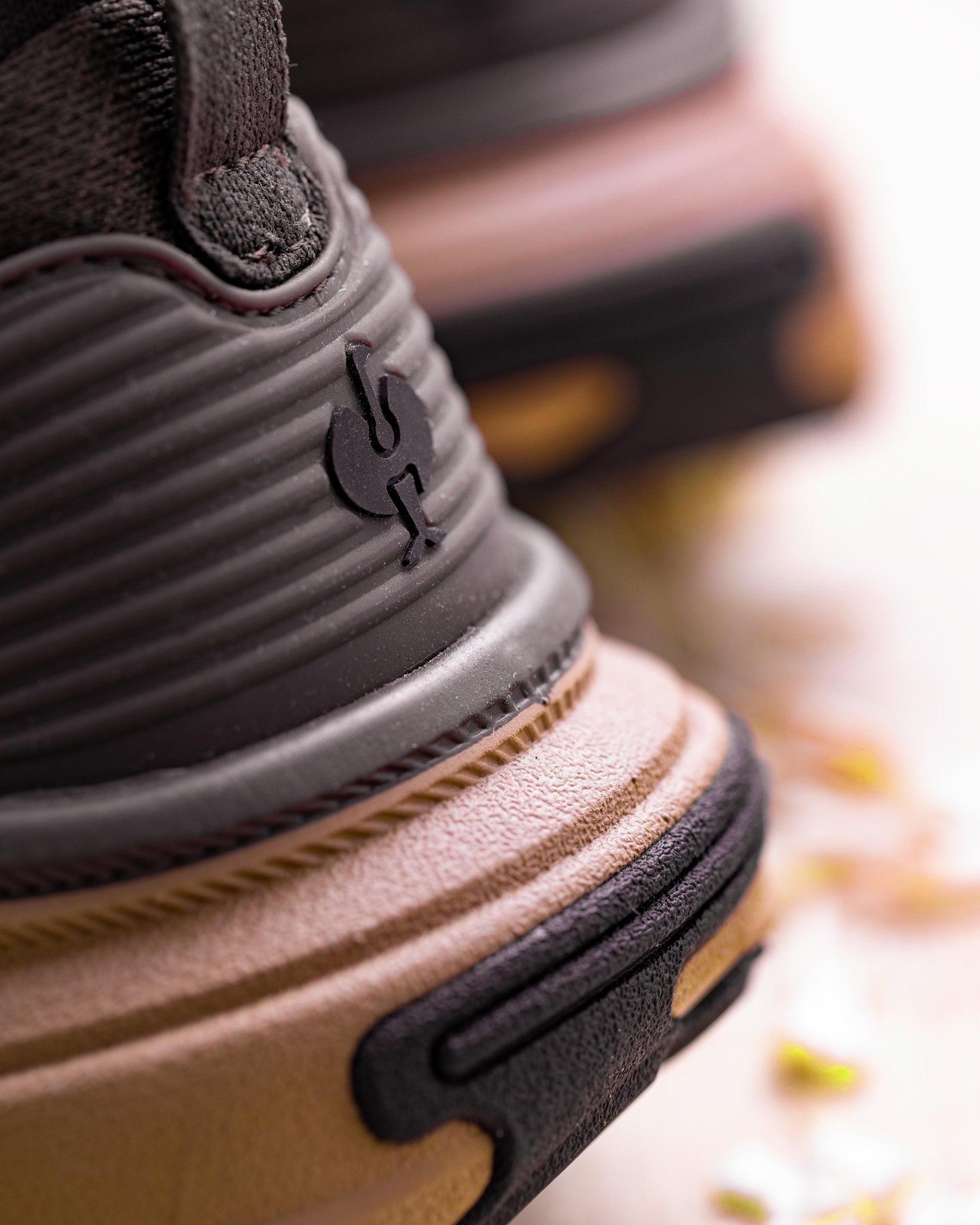 Close-up of a chunky sneaker showing the heel and side profile, featuring a dark gray ribbed collar with a black logo, a tan textured midsole, and a black outsole.