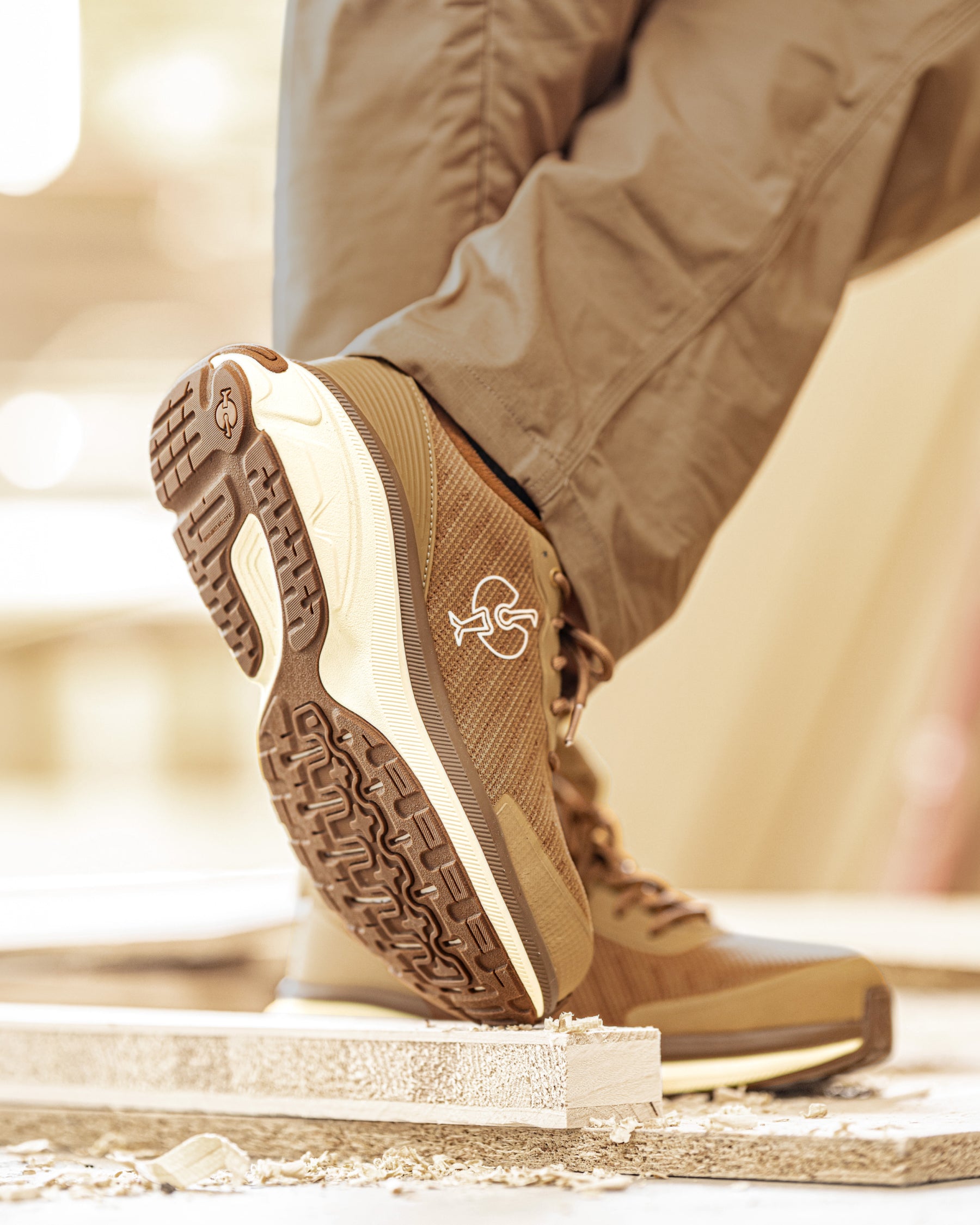 A close-up lifestyle shot of a person's legs and feet wearing brown and beige athletic sneakers, with one shoe lifted to show the sole and tread pattern. The person is also wearing khaki pants, and the background is a softly blurred warm-toned indoor setting.