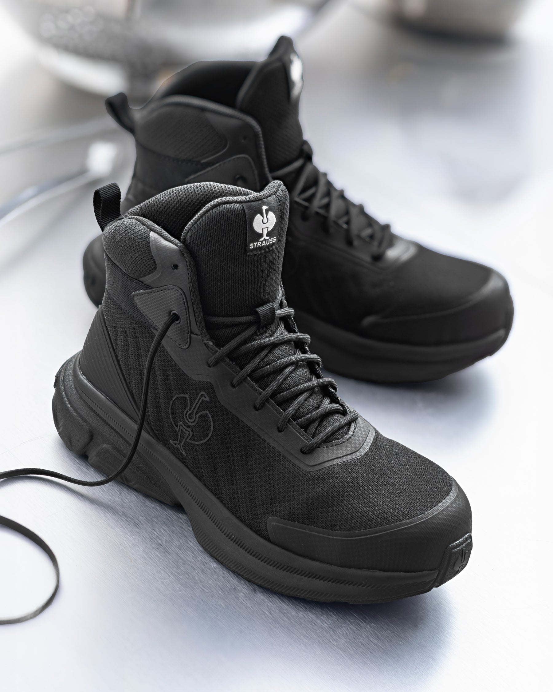 A pair of black high-top tactical boots with textured mesh panels, black laces, and a white logo on the tongue, displayed on a reflective metallic surface.