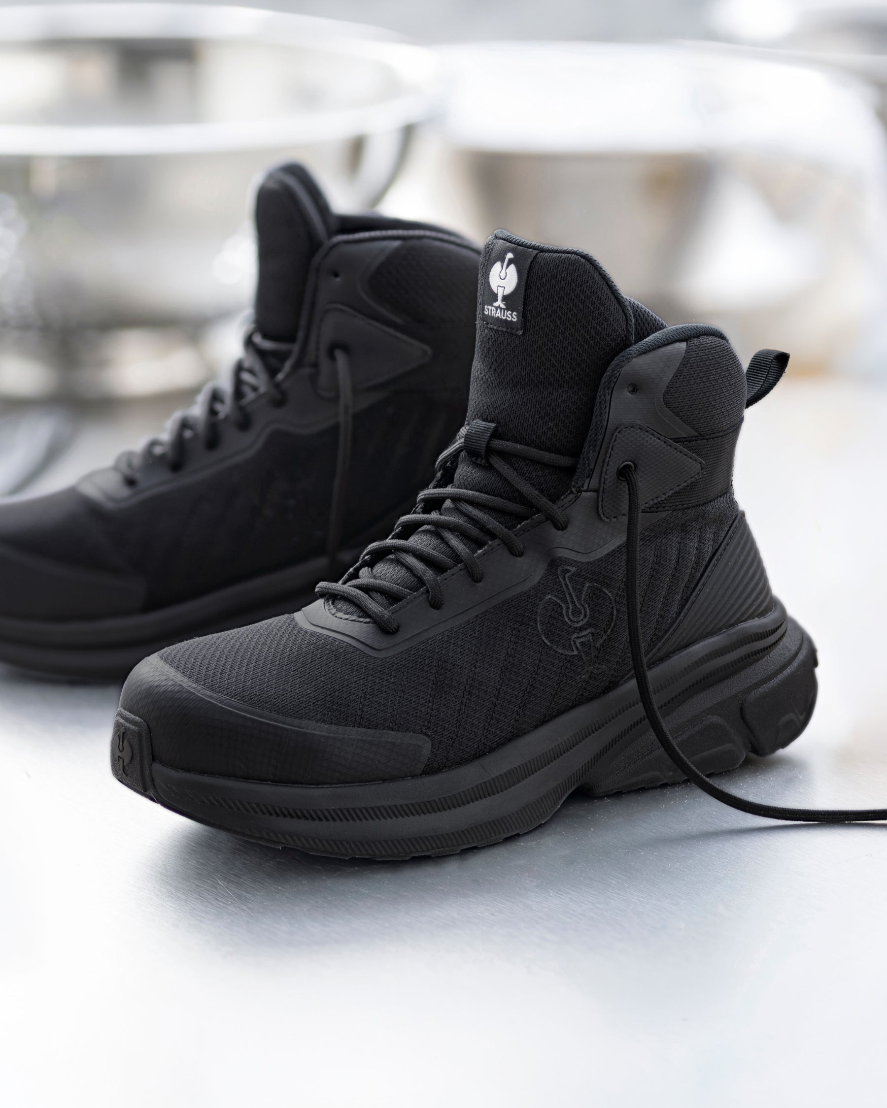 A pair of black high-top tactical boots with matching black laces and soles, displayed on a reflective light gray surface with a blurred studio background.
