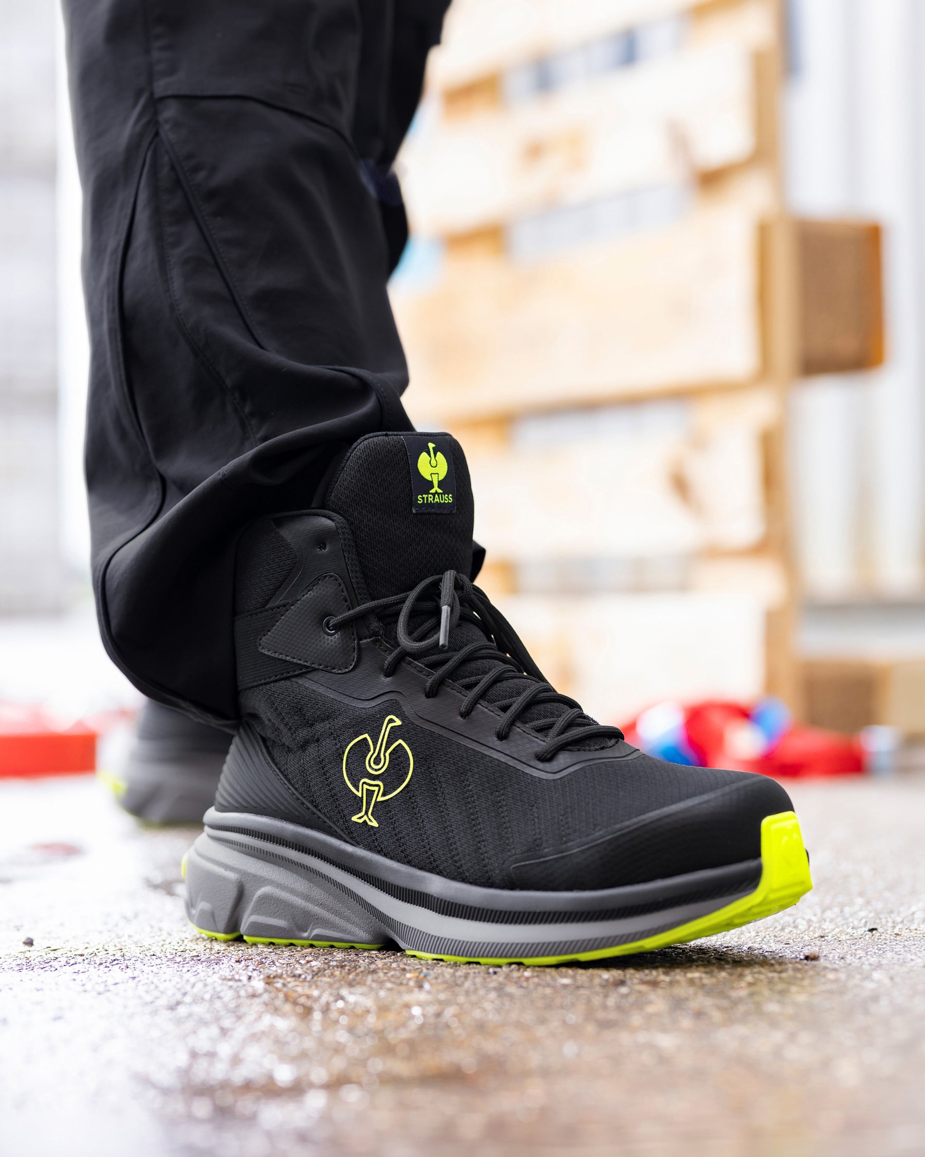 A close-up of a person's foot wearing a black high-top work boot with a bright neon yellow-green logo and sole accent, paired with black pants.