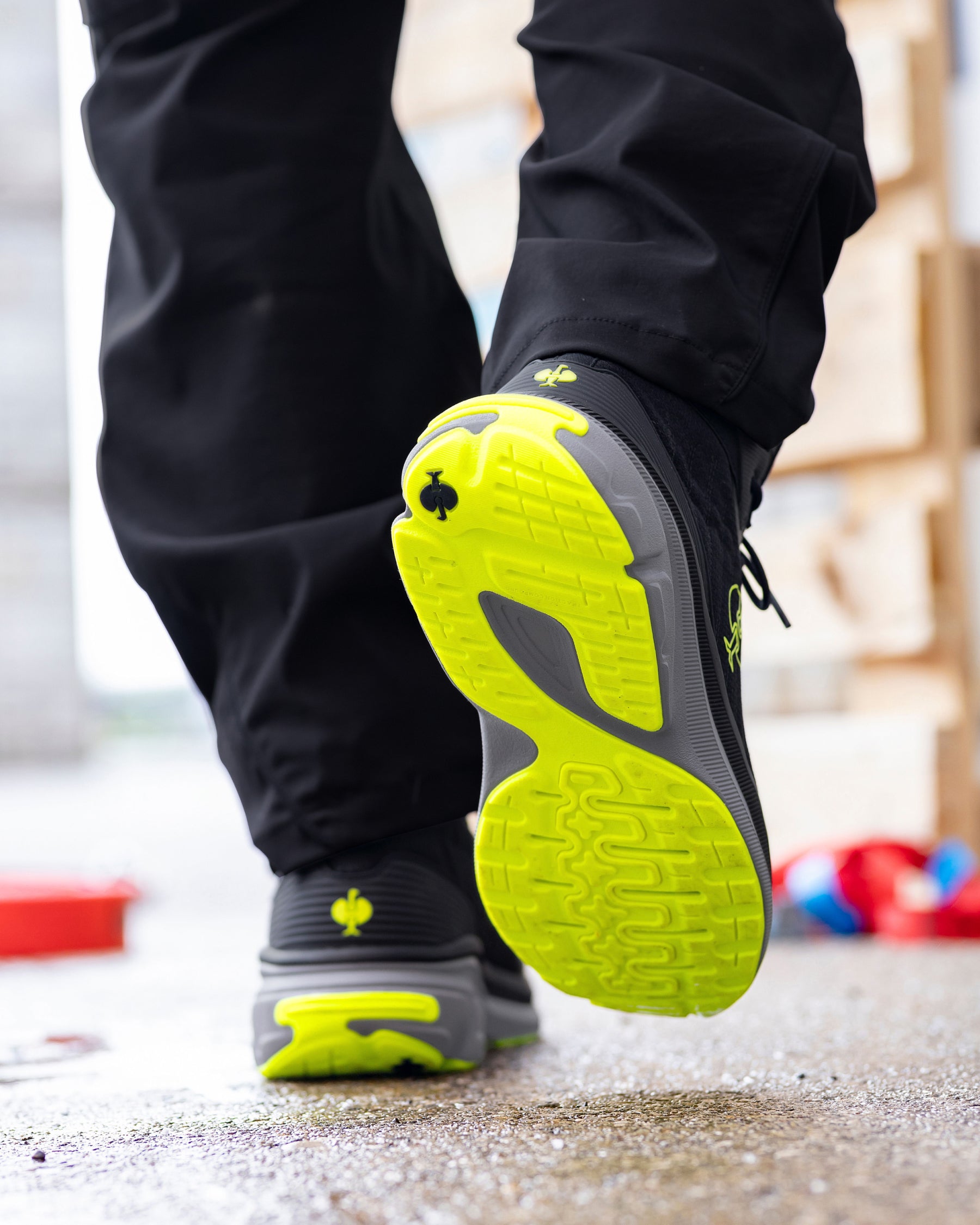 A close-up of a person's legs wearing black pants and black athletic sneakers with bright neon yellow-green soles, shown walking on a concrete surface.