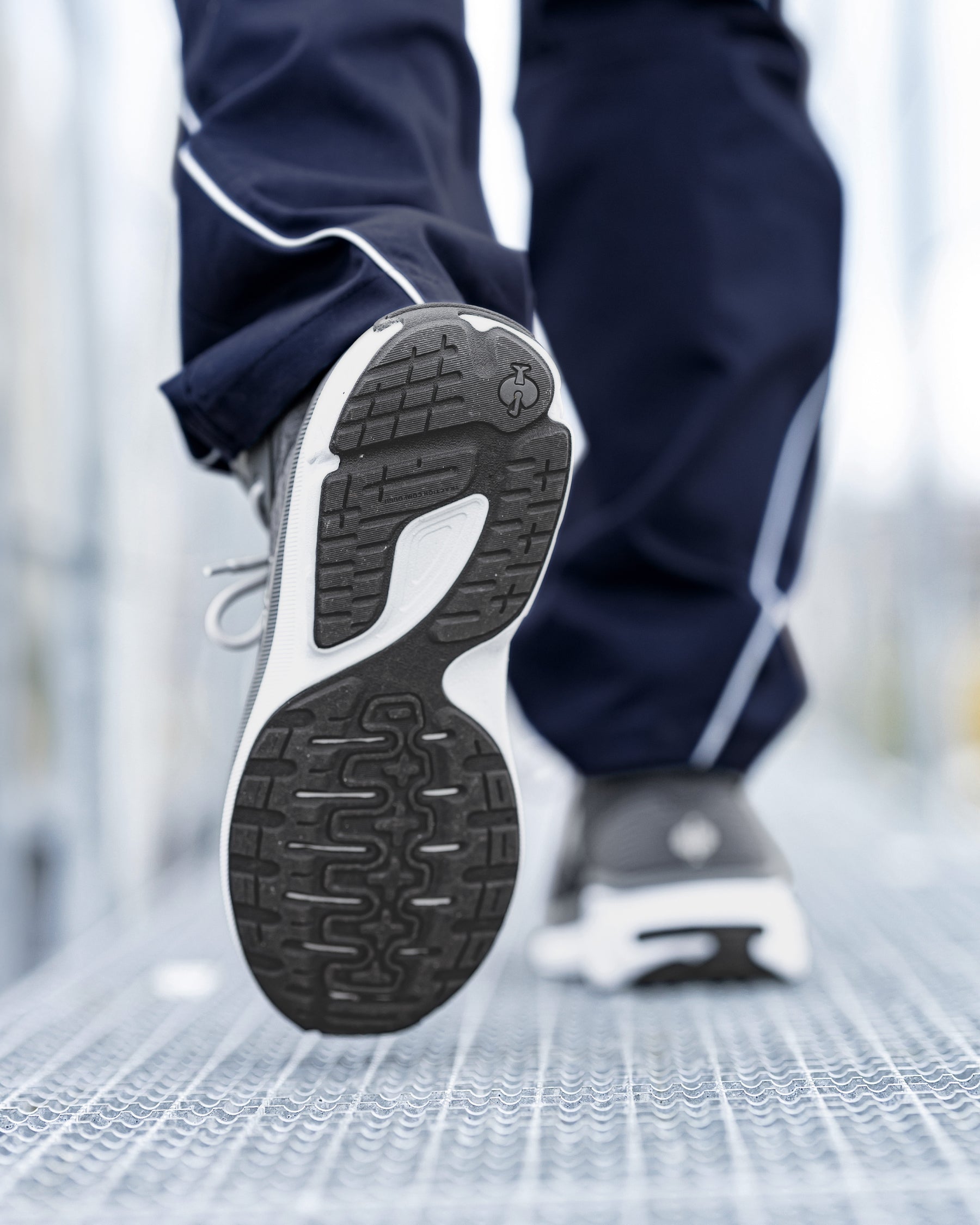 A close-up action shot of a person's leg lifting a black and white athletic sneaker off a metal walkway, with navy blue track pants visible in the background.