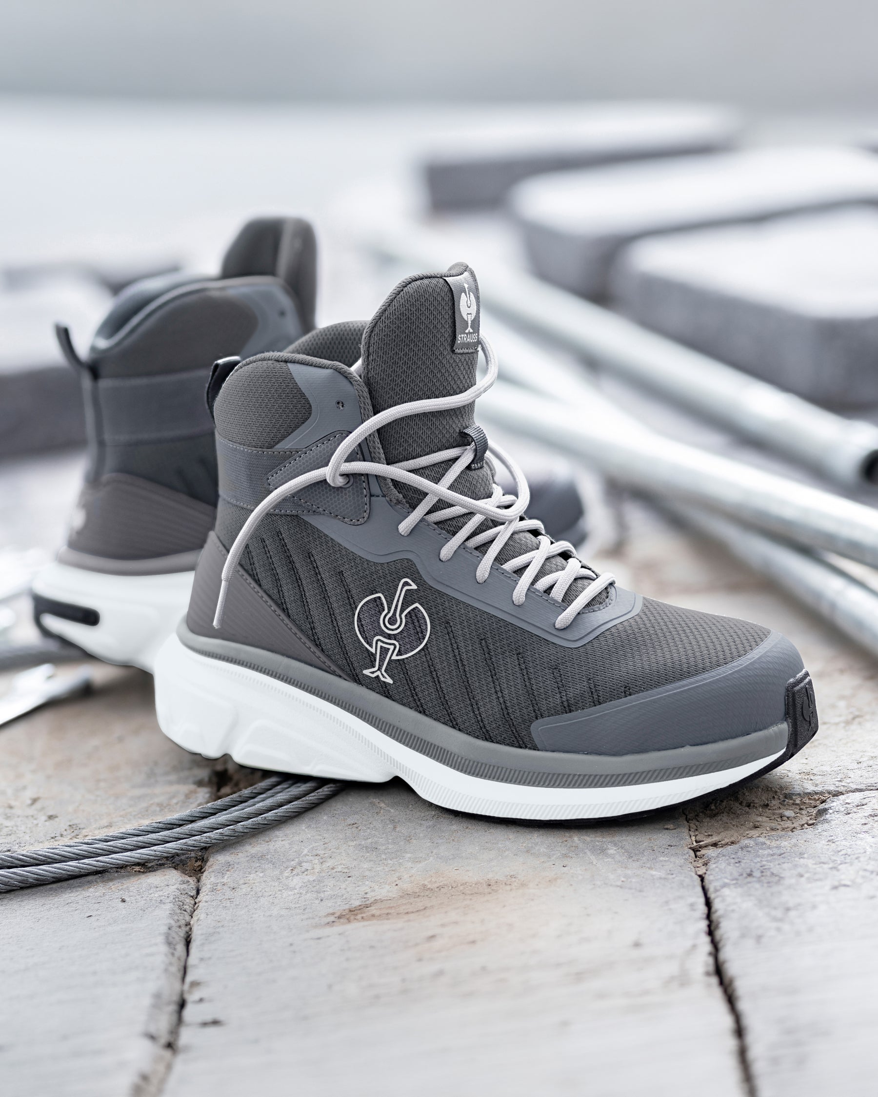 A pair of mid-top athletic sneakers in dark gray and white, featuring a textured mesh upper, white laces, and a white sole, displayed on a weathered wooden surface with a blurred industrial background.