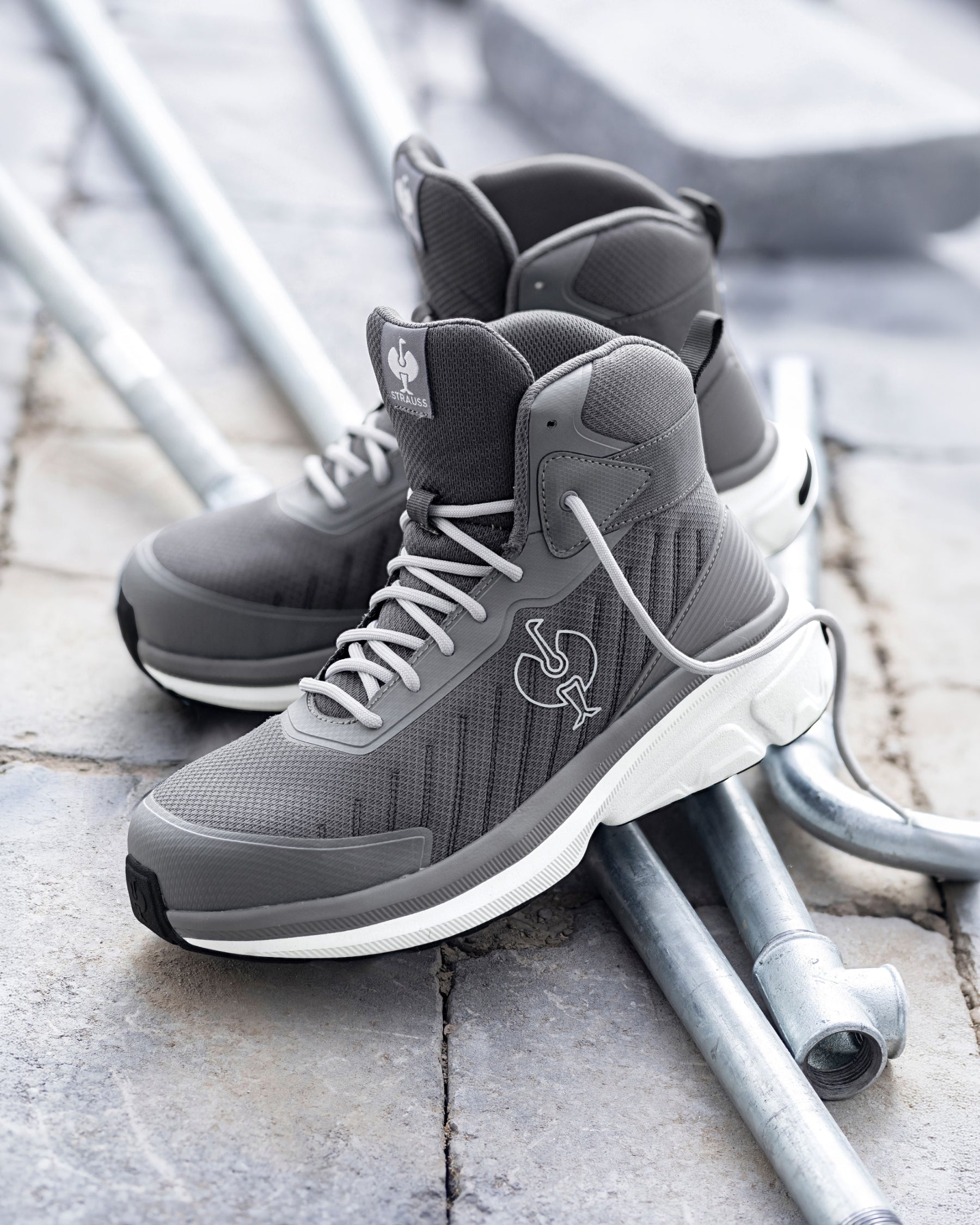 A pair of gray high-top sneakers with white midsoles and a white Spurs logo, resting on metal pipes over a stone pavement background.