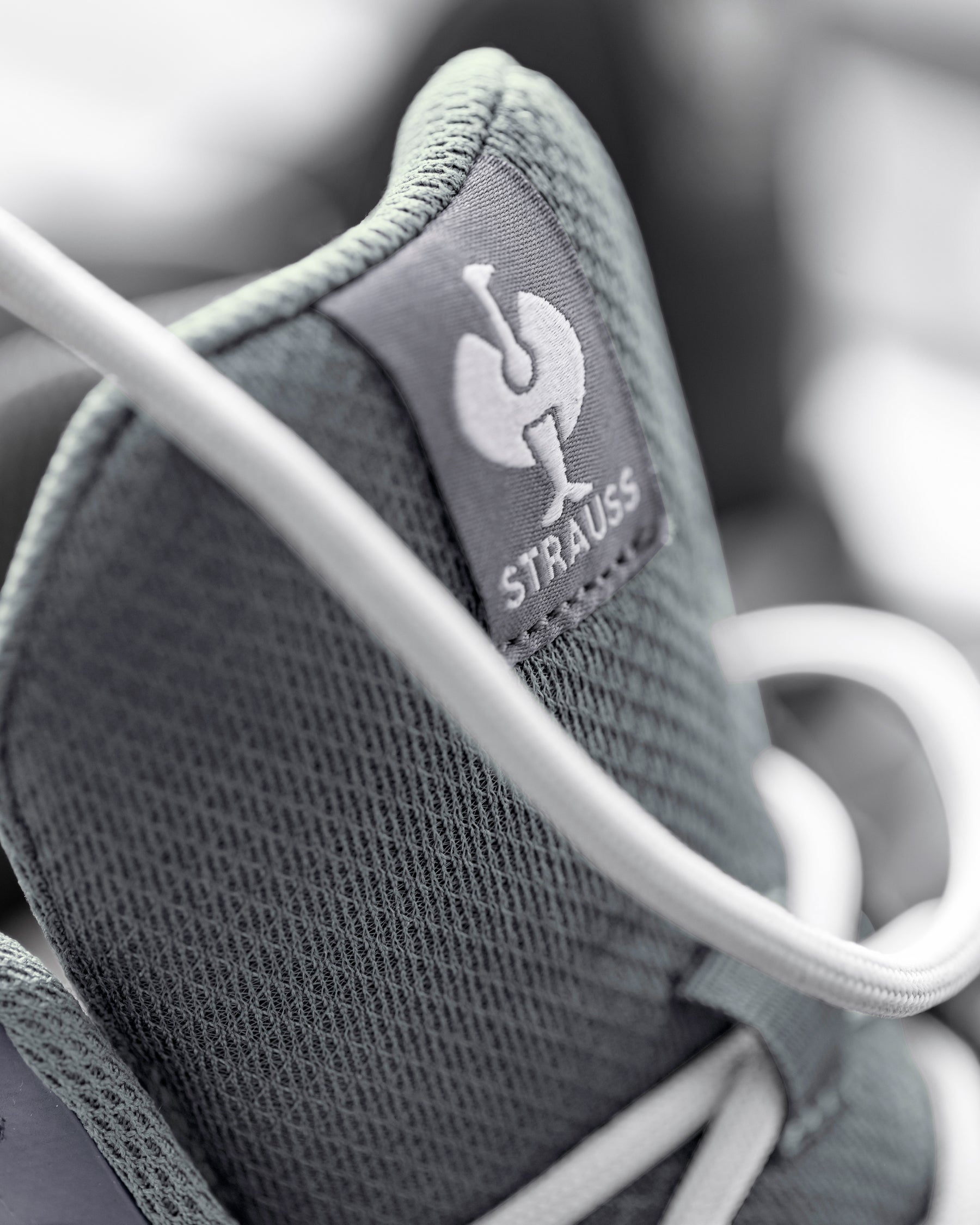 Close-up of a gray mesh sneaker with white laces, showing a gray tongue label with the white Strauss logo and brand name.